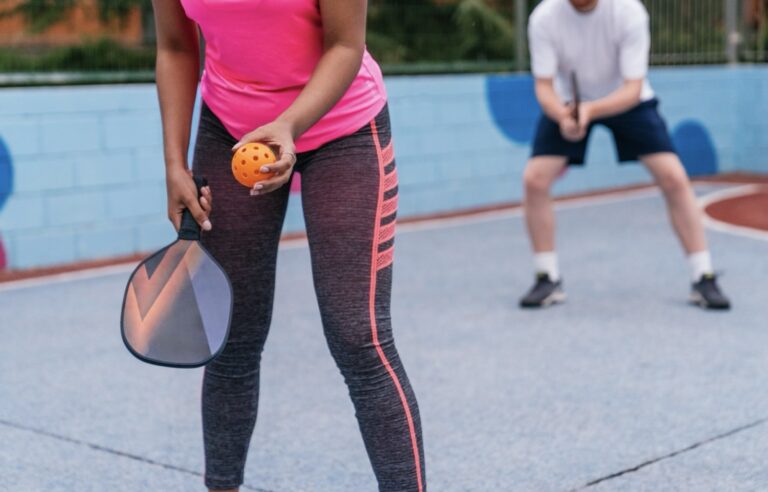 Woman holding a pickleball paddle and ball, ready to serve while her partner waits across the court