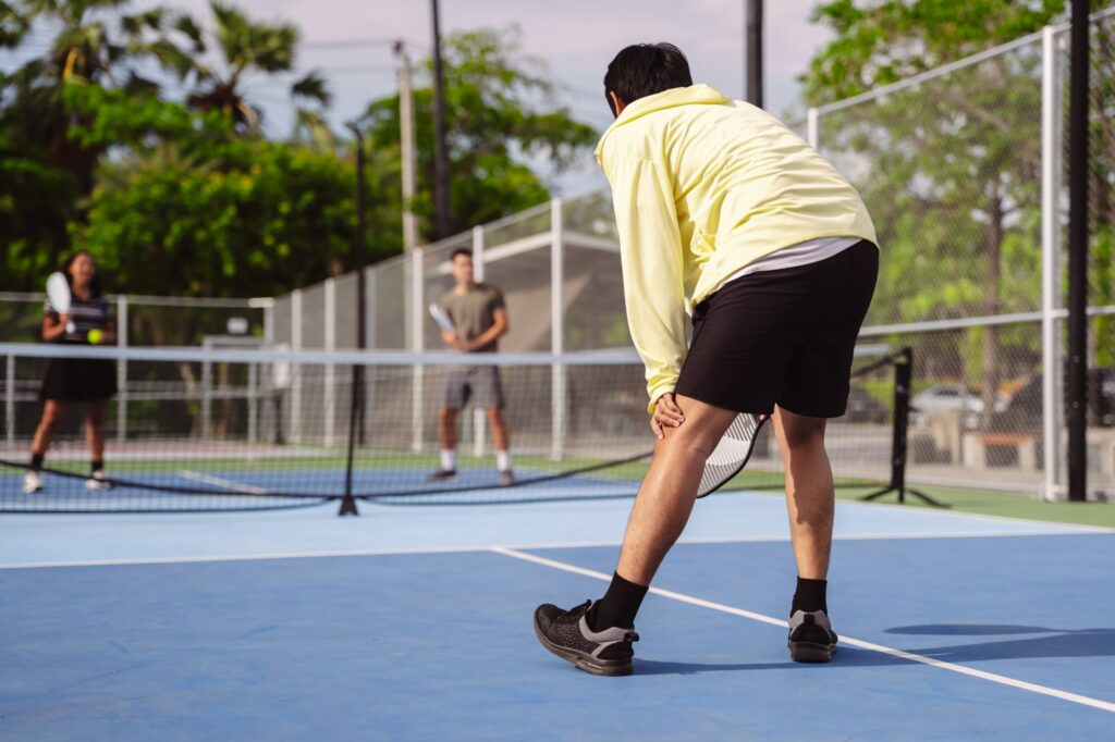 A pickleball player waiting in ready position at the baseline during a doubles match